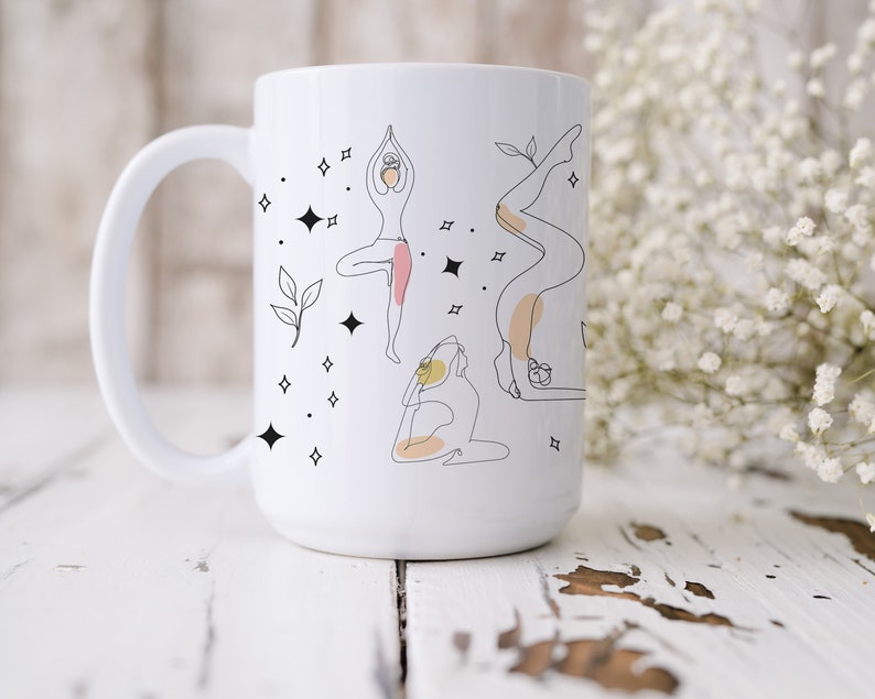 May include: White ceramic mug with line art illustrations of yoga poses in black, with pink and peach accents. The design includes stars and leaves. The mug has a large handle and is set against a white wooden surface.