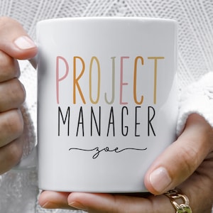 Könnte beinhalten: Weiße Keramiktasse mit dem Text "PROJECT MANAGER" in bunten Buchstaben und dem Namen "zoe" darunter in Kursivschrift.