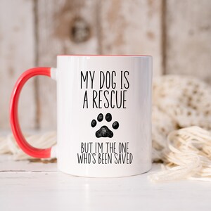 May include: White ceramic coffee mug with a red rim and handle. The mug has black text that reads "My dog is a rescue but I'm the one who's been saved" with a black paw print.