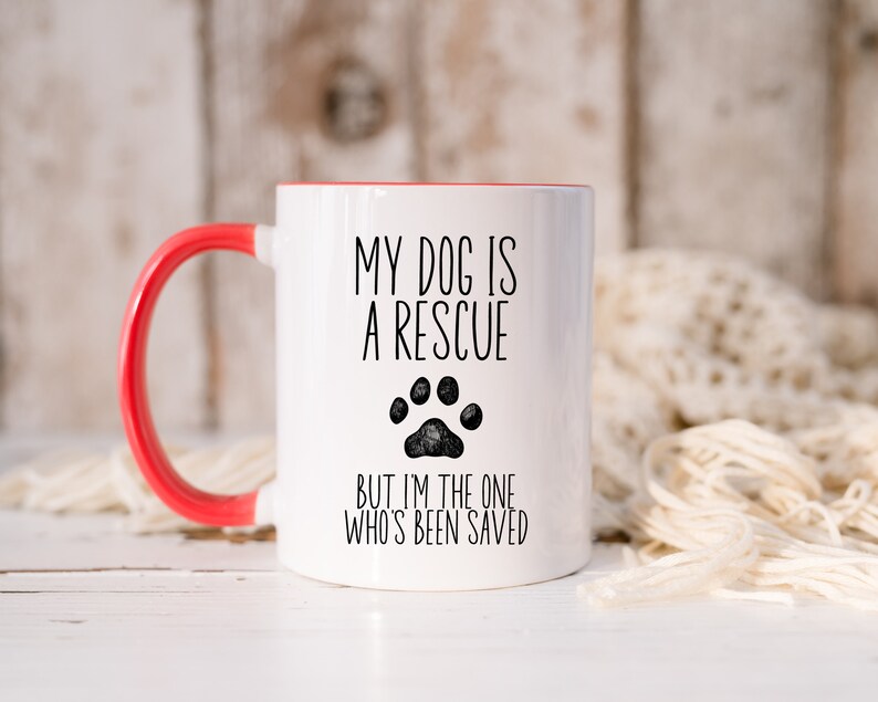 May include: White ceramic coffee mug with a red rim and handle. The mug has black text that reads "My dog is a rescue but I'm the one who's been saved" with a black paw print.