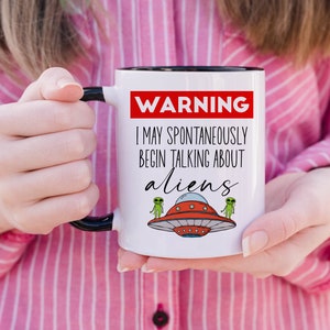 Op de afbeelding: Een witte keramische mok met een zwarte rand en handvat. De mok heeft een rood waarschuwingsbord met de tekst "WAARSCHUWING IK KAN SPONTAN BEGINNEN TE PRATEN OVER BUITENAARDSE WEZENS". Er zijn twee groene buitenaardse wezens en een vliegende schotel op de mok.