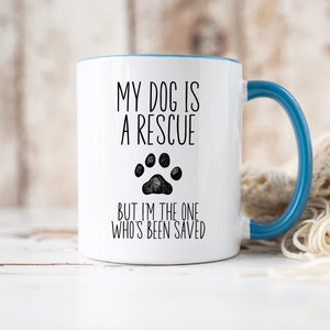 May include: White ceramic coffee mug with a blue rim and handle. The mug has black text that reads "My dog is a rescue but I'm the one who's been saved" with a black paw print graphic.