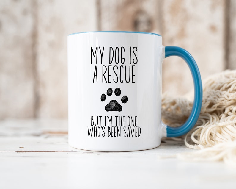May include: White ceramic coffee mug with a blue rim and handle. The mug has black text that reads "My dog is a rescue but I'm the one who's been saved" with a black paw print graphic.
