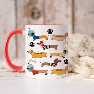 May include: White ceramic coffee mug with a red rim and handle. The mug features a repeating pattern of cartoon dachshunds wearing different costumes, including a hot dog, a sailor, and an astronaut.