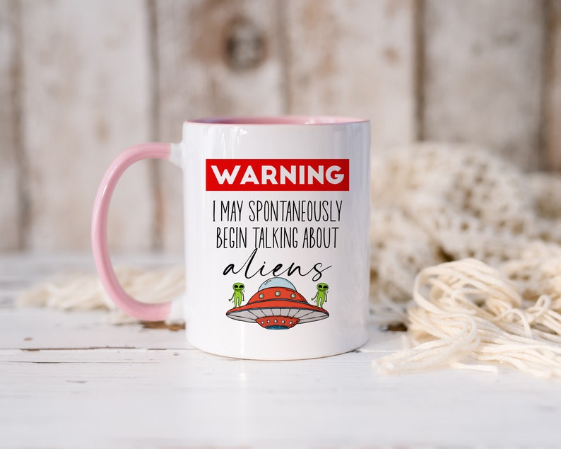 Op de afbeelding: Witte keramische mok met een roze handvat. De mok heeft een rood waarschuwingsbord met de tekst "WAARSCHUWING IK KAN SPONTAN BEGINNEN TE PRATEN OVER BUITENAARDSE WEZENS". Er zijn twee groene buitenaardse wezens in vliegende schotels op de mok.