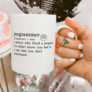 May include: White ceramic mug with a black and white definition of the word "programmer" printed on it. The definition reads: "programmer. noun. a person who fixed a program you didn't know you had in a way that you don't understand."