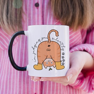 May include: White ceramic mug with a black handle. The mug features a cartoon illustration of a cat with its back turned, wearing yellow boots and a green plant growing from its head. The text "I'm judging you silently" is written on the mug.