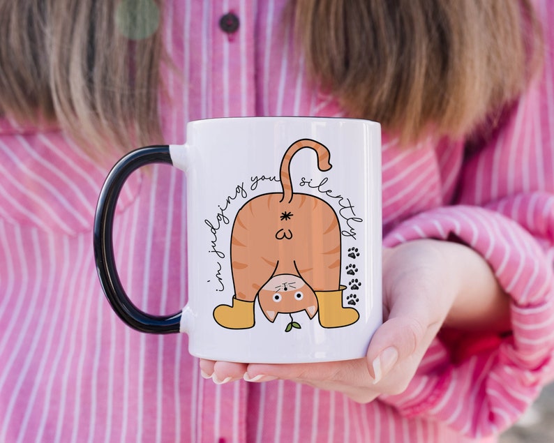 May include: White ceramic mug with a black handle. The mug features a cartoon illustration of a cat with its back turned, wearing yellow boots and a green plant growing from its head. The text "I'm judging you silently" is written on the mug.