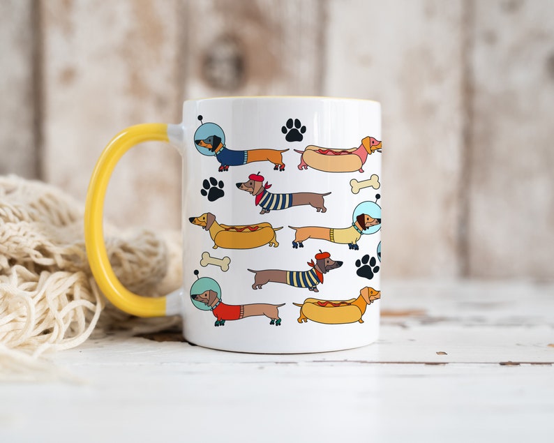 May include: White ceramic mug with a yellow handle. The mug features a repeating pattern of cartoon dachshunds wearing different outfits and accessories, including a hot dog costume, a beret, and a space helmet.