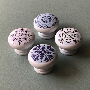 May include: Four white ceramic knobs with blue and purple patterned designs. The knobs are round and have a raised edge around the top.