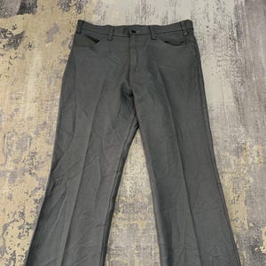 Puede incluir: Pantalones grises con un diseño clásico de pierna recta. Los pantalones tienen cierre de botón y cremallera, trabillas para el cinturón y bolsillos delanteros y traseros. La tela parece ser de peso medio.