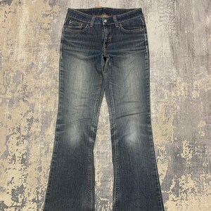 Op de afbeelding: Blauwe spijkerbroek met een klassiek ontwerp met vijf zakken en een lichte flare aan de zoom. De jeans heeft een vervaagde wassing en een knoop- en ritssluiting. De stof lijkt versleten, wat wijst op een vintage of tweedehands item.