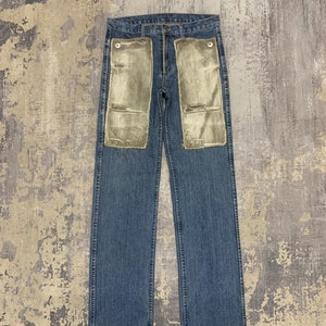 May include: Blue denim jeans with two large, light green velvet panels sewn onto the front. The jeans have a classic five-pocket design and a button and zipper closure. The velvet panels are rectangular and extend from the waist to the mid-thigh.
