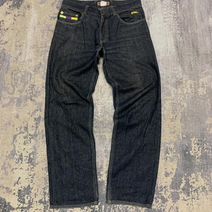 May include: Dark indigo denim jeans featuring a classic five-pocket design with contrast stitching. The right front pocket displays a small rectangular patch with yellow and purple stripes, while the left pocket features a small yellow embroidered logo.
