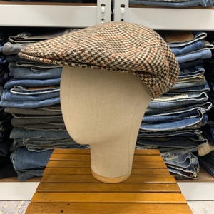 May include: A brown and tan tweed flat cap with a classic houndstooth pattern. The cap is displayed on a mannequin head, showcasing its shape and design. The hat is made of a textured fabric and has a short peak.