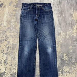 May include: Dark wash denim jeans with a classic five-pocket design. The jeans have a straight leg cut and show signs of wear, including a small tear on the right knee. The denim has faded areas, giving the jeans a vintage look.