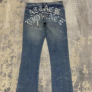 May include: Faded blue denim jeans with a bootcut leg. The back pockets feature white gothic lettering that reads "ATTACK WORK". The jeans have a distressed, textured appearance.