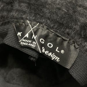 May include: Close-up of a black Kangol design hat. The hat has a fuzzy texture and a black band. A black label inside the hat reads "KANGOL design" and "MADE IN ENGLAND".
