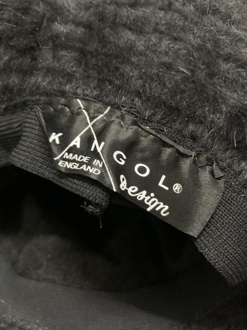 May include: Close-up of a black Kangol design hat. The hat has a fuzzy texture and a black band. A black label inside the hat reads "KANGOL design" and "MADE IN ENGLAND".