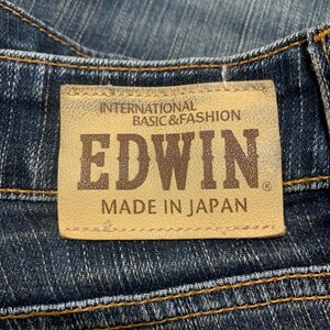 Op de afbeelding: Close-up van een jeanslabel. Het label is bruin met de merknaam "EDWIN" in bruine letters. De tekst "INTERNATIONAL BASIC & FASHION" en "MADE IN JAPAN" zijn ook zichtbaar.