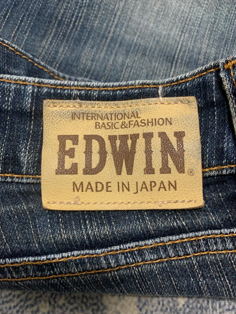 Op de afbeelding: Close-up van een jeanslabel. Het label is bruin met de merknaam "EDWIN" in bruine letters. De tekst "INTERNATIONAL BASIC & FASHION" en "MADE IN JAPAN" zijn ook zichtbaar.