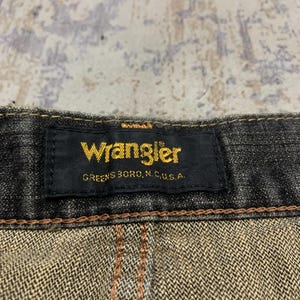Pu&ograve; includere: Primo piano di un'etichetta nera di jeans Wrangler con scritte dorate. L'etichetta recita "Wrangler" e "Greensboro, N.C., U.S.A." I jeans hanno cuciture marroni e un aspetto denim strutturato.