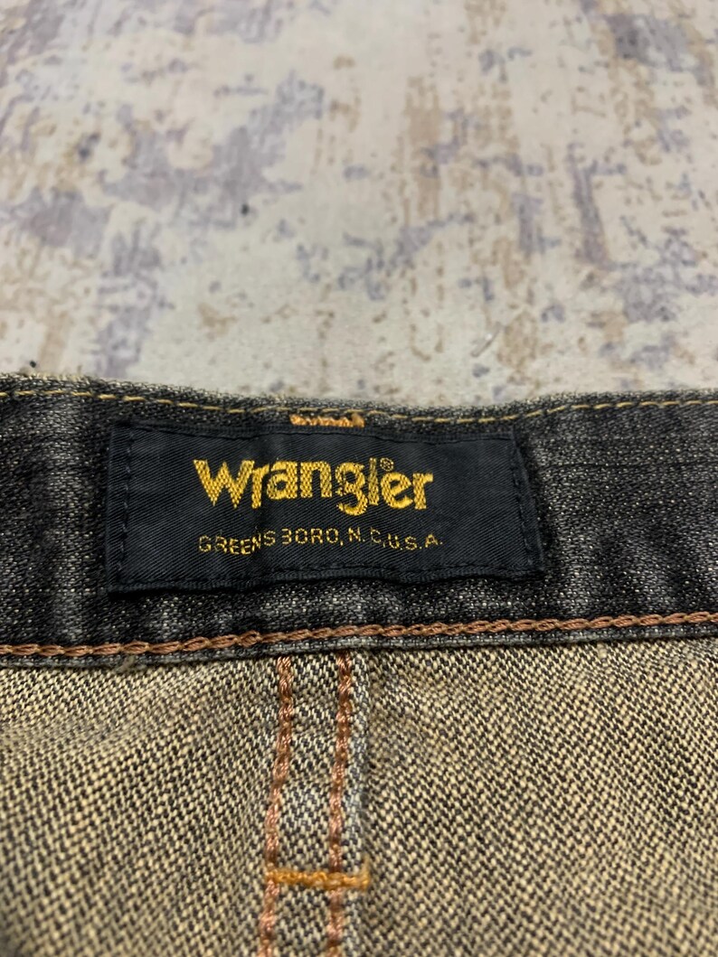 Pu&ograve; includere: Primo piano di un'etichetta nera di jeans Wrangler con scritte dorate. L'etichetta recita "Wrangler" e "Greensboro, N.C., U.S.A." I jeans hanno cuciture marroni e un aspetto denim strutturato.