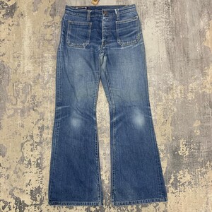 May include: Blue denim jeans featuring a high-waisted fit, a button-fly closure, and two prominent square patch pockets on the front. The legs have a distinct flared silhouette with faded areas on the thighs and knees, finished with a hem.