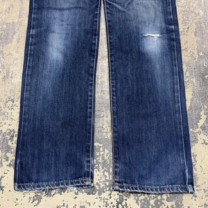 May include: Close-up of a pair of blue denim jeans. The jeans have a classic five-pocket design, with a straight leg cut. The denim has a faded wash with some wear and tear, including a small rip on the right leg.