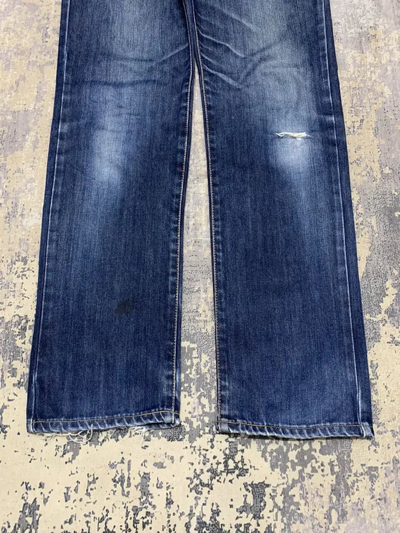 May include: Close-up of a pair of blue denim jeans. The jeans have a classic five-pocket design, with a straight leg cut. The denim has a faded wash with some wear and tear, including a small rip on the right leg.