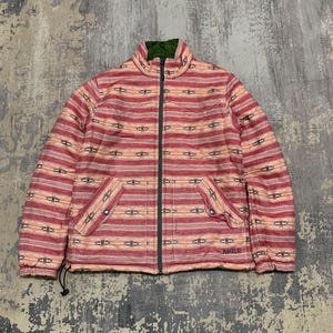 May include: A red and peach-coloured jacket with a geometric pattern. The jacket has a full zip, two front pockets, and a green interior collar. The brand name "AIGLE" is visible on the lower right side.