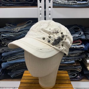 May include: A white cotton baseball cap with a skull and star embellishments. The cap has a distressed look and is decorated with metal studs and charms.