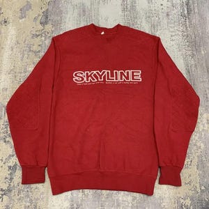 May include: Red crew neck sweatshirt featuring the word "SKYLINE" in white. The sweatshirt has quilted elbow patches and the text "Love a fresh and new everyday. Skyline is car with a loving new spirit."