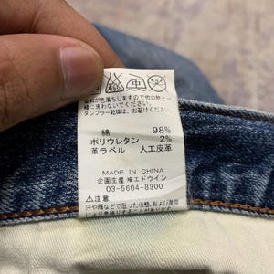 May include: Close-up of a denim jeans label with Japanese text and care symbols. The label indicates the jeans are made in China, with 98% cotton and 2% polyurethane. The label also includes washing instructions.