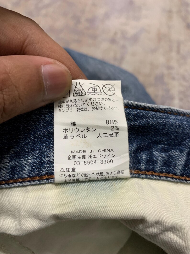 May include: Close-up of a denim jeans label with Japanese text and care symbols. The label indicates the jeans are made in China, with 98% cotton and 2% polyurethane. The label also includes washing instructions.