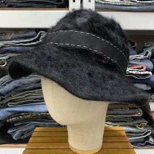May include: A black, fuzzy bucket hat with a wide, black band featuring white stitching. The hat is displayed on a mannequin head, showcasing its shape and texture. The hat's brim is slightly curved.