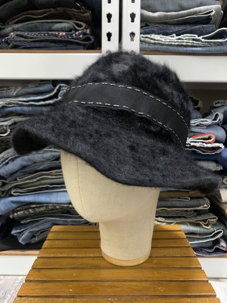 May include: A black, fuzzy bucket hat with a wide, black band featuring white stitching. The hat is displayed on a mannequin head, showcasing its shape and texture. The hat's brim is slightly curved.