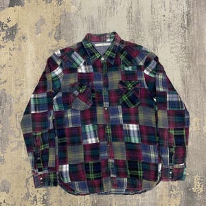 May include: A long-sleeved patchwork plaid shirt in shades of green, purple, blue, and white. The shirt features a button-down front, two chest pockets, and buttoned cuffs. The fabric has a soft, worn appearance.