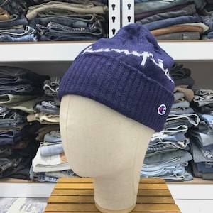 May include: A navy blue knit beanie with a white mountain range design across the front. The beanie features a small red and white "C" logo on the side. The hat is displayed on a mannequin head.