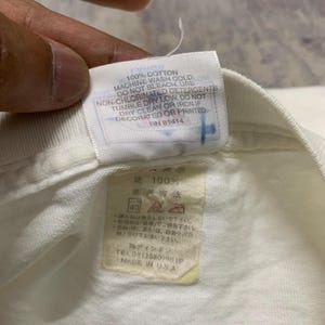 Puede incluir: Primer plano de una etiqueta de prenda de algod&oacute;n blanco con texto en ingl&eacute;s y japon&eacute;s. La etiqueta incluye instrucciones de lavado y el texto "MADE IN USA".