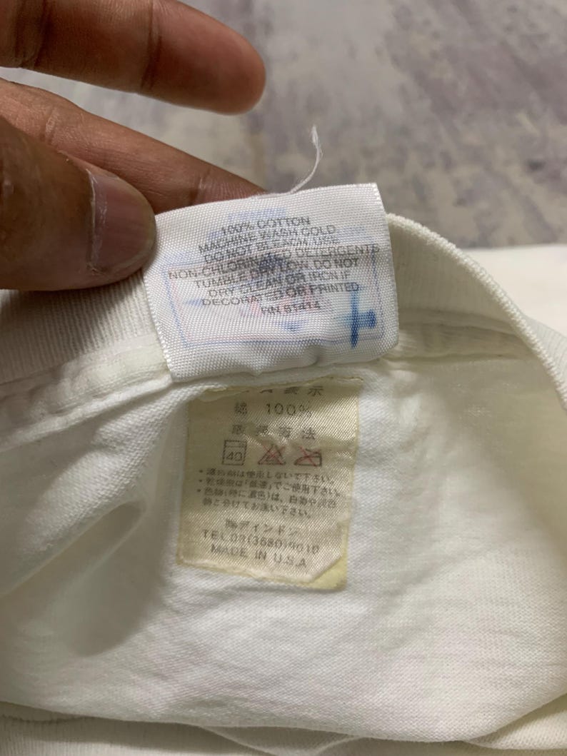 Puede incluir: Primer plano de una etiqueta de prenda de algod&oacute;n blanco con texto en ingl&eacute;s y japon&eacute;s. La etiqueta incluye instrucciones de lavado y el texto "MADE IN USA".