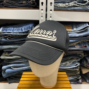 May include: A black trucker cap featuring a mesh back and a curved brim with a white cord detail. The front panel displays the word Kauai in a stylized cream-coloured script font with a wave-like graphic underline, centred on the crown.