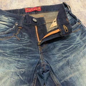 May include: Close-up of blue denim jeans with an orange zipper and stitching. The jeans have a faded, distressed look with visible wear patterns. A red tag is visible on the waistband.