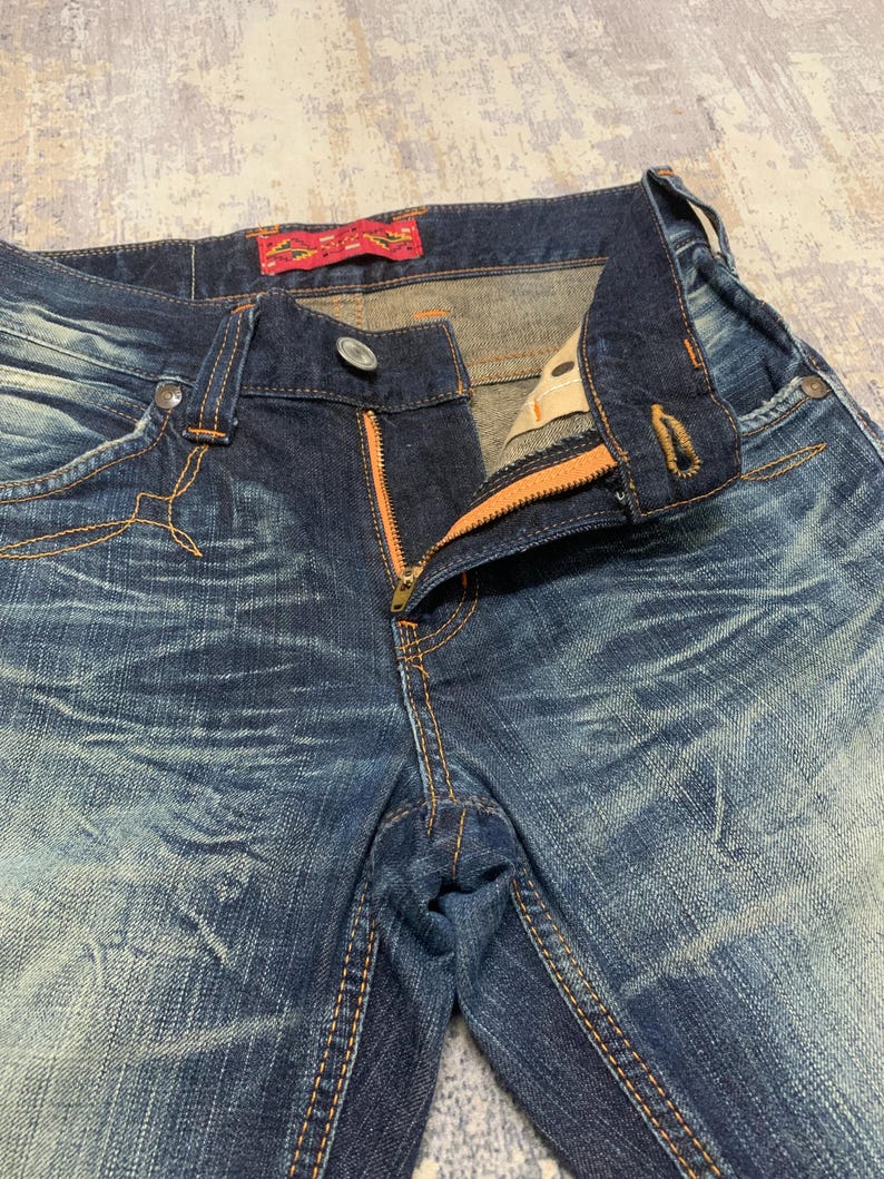May include: Close-up of blue denim jeans with an orange zipper and stitching. The jeans have a faded, distressed look with visible wear patterns. A red tag is visible on the waistband.