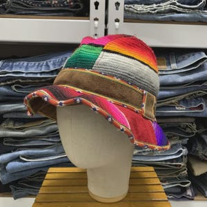 Pode incluir: Um chapéu bucket com um design de retalhos de tecido às riscas multicoloridas em tons de vermelho, verde, laranja e cinzento. O chapéu inclui uma faixa de camurça castanha à volta da copa e um rebordo decorativo com um padrão geométrico.