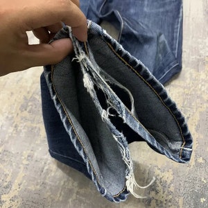 May include: Close-up of blue denim jeans with frayed edges. The jeans are held open, showcasing the inside seam and the distressed detailing. The denim has a classic wash and visible stitching.