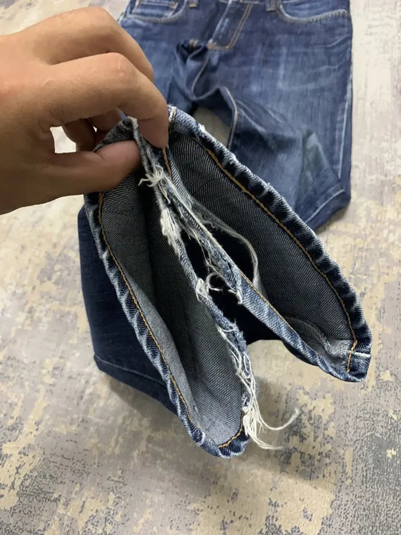 May include: Close-up of blue denim jeans with frayed edges. The jeans are held open, showcasing the inside seam and the distressed detailing. The denim has a classic wash and visible stitching.