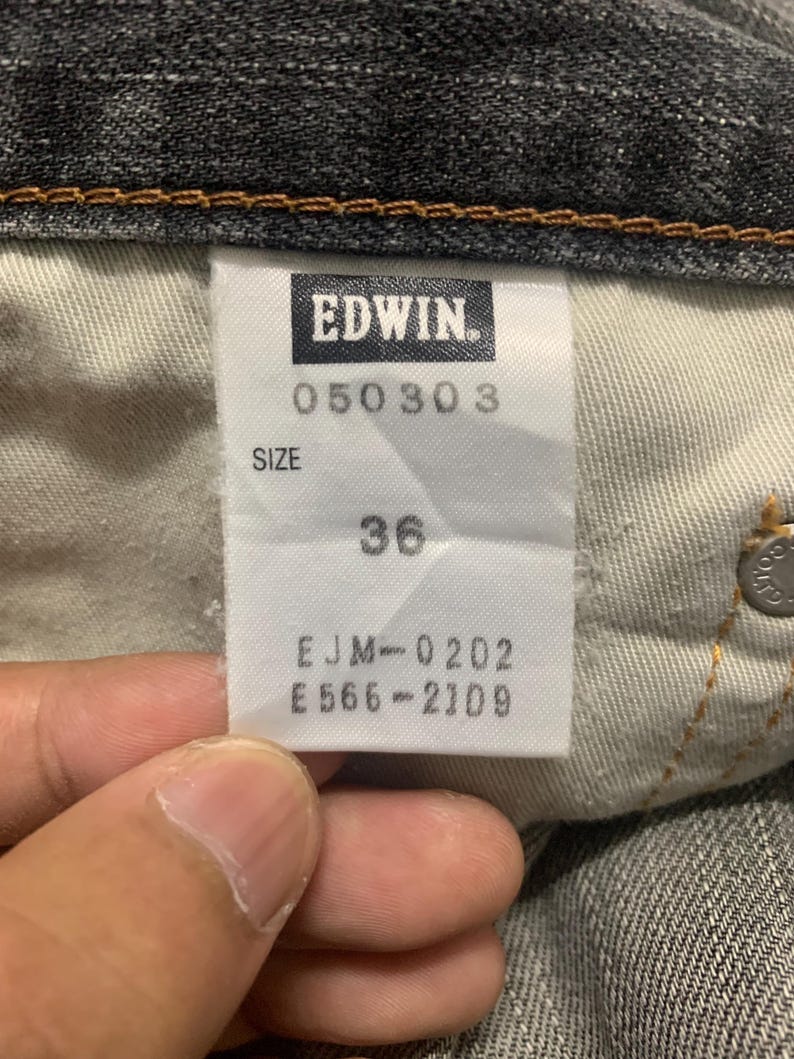 Pode incluir: Close-up de uma etiqueta de jeans Edwin. A etiqueta branca apresenta texto preto, incluindo a marca, c&oacute;digos de produto e tamanho 36. Os jeans s&atilde;o de cor cinza escuro.