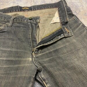 Pu&ograve; includere: Primo piano di un paio di jeans Wrangler grigio scuro con la cerniera parzialmente aperta. I jeans hanno un classico design a cinque tasche, con cuciture visibili e chiusura con bottone. Il logo Wrangler &egrave; visibile sulla cintura.
