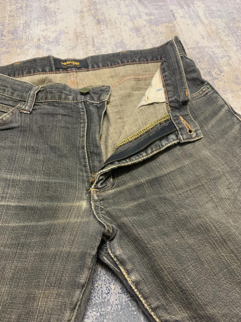 Pu&ograve; includere: Primo piano di un paio di jeans Wrangler grigio scuro con la cerniera parzialmente aperta. I jeans hanno un classico design a cinque tasche, con cuciture visibili e chiusura con bottone. Il logo Wrangler &egrave; visibile sulla cintura.